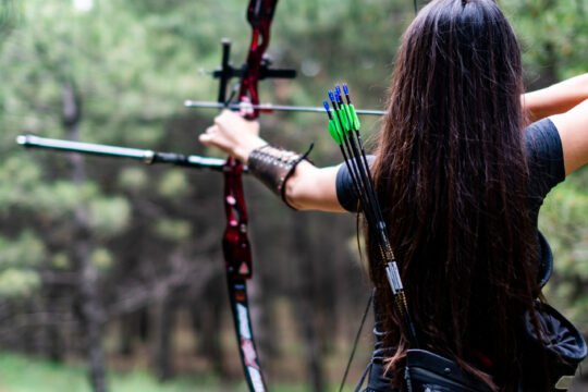 An athletic female aiming with bow and arrow towards the trees
